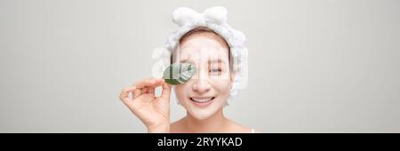 Banner der hübschen Frau hält grünes Blatt, schaut nachdenklich zur Seite, bringt Tonmaske auf das Gesicht, Stockfoto