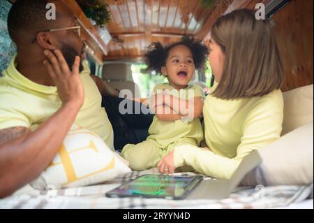 Auf der Rückseite eines weißen klassischen Minibusses spielen kleine Kinder und erfahren auf ihrem Tablet mehr über die Welt, während sich die Eltern um Hilfe kümmern. Stockfoto