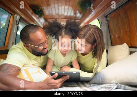 Auf der Rückseite eines weißen klassischen Minibusses spielen kleine Kinder und erfahren auf ihrem Tablet mehr über die Welt, während sich die Eltern um Hilfe kümmern. Stockfoto