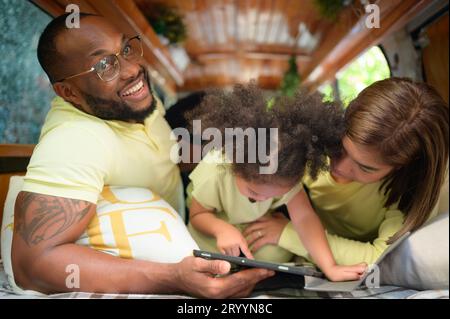 Auf der Rückseite eines weißen klassischen Minibusses spielen kleine Kinder und erfahren auf ihrem Tablet mehr über die Welt, während sich die Eltern um Hilfe kümmern. Stockfoto