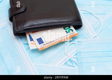 Schwarze Herren Brieftasche mit Euro Geld innen auf den blauen Einweg-medizinischen Gesichtsmasken. Handel mit medizinischen Masken. Unternehmen während einer Pandemie oder Epidemie Stockfoto