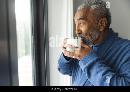 Rücksichtsvoller älterer, birassistischer Mann, der Bademantel trägt und zu Hause Kaffee am Fenster trinkt Stockfoto