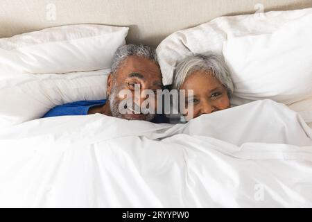 Porträt eines glücklichen älteren, birassischen Paares, das zu Hause im Bett lag und mit Bettwäsche bedeckt war Stockfoto