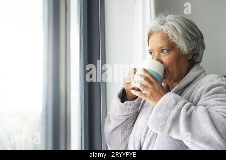 Rücksichtsvolle ältere, birassische Frau, die Bademantel trägt und zu Hause Kaffee am Fenster trinkt Stockfoto