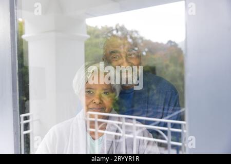 Glückliches älteres birassisches Paar, das Bademäntel trägt und zu Hause durch das Fenster schaut Stockfoto