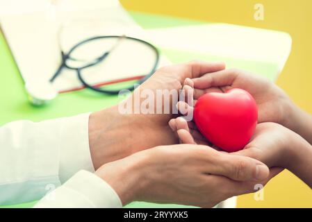 Arzthände halten und geben dem Patienten kleine Kinder ein rotes Massageherz, um sich von der Krankheit zu erholen. Krankenhaus und Gesundheitswesen Stockfoto