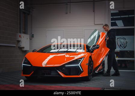 Ein orangefarbener Lamborghini Revuelto steht mit geöffneter Scherentür im Innenbereich und zeigt sein elegantes Design. Eine Person steht daneben auf einem karierten flo Stockfoto