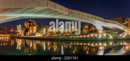 Dies ist ein nächtlicher Blick auf das Guggenheim Museum in Bilbao, das seine ikonische Architektur, die Alfonso-Brücke und die Reflexionen auf dem Fluss zeigt. Stockfoto