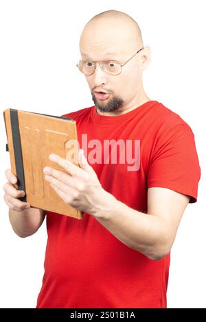 Ein überraschter Mann, der ein rotes T-Shirt und eine Brille trägt, hält ein Tablet in beiden Händen. Sein Gesichtsausdruck zeigt Erstaunen, wenn er auf den Geröll schaut Stockfoto