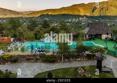 Ein großer Pool ist von einem üppigen grünen Rasen und einem Steinweg umgeben. Der Pool ist von einer Gruppe von Leuten umgeben, von denen einige auf den Stühlen A sitzen Stockfoto