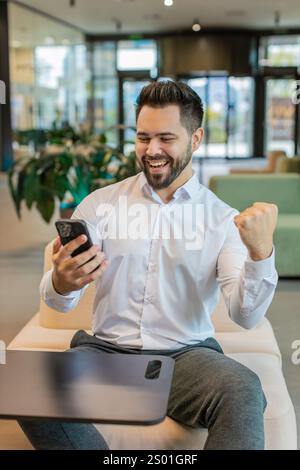 Glücklicher junger Geschäftsmann in weißem Hemd, der am Smartphone arbeitet, schockiert von einem plötzlichen Siegesspiel, das Lotterie-Jackpot-Leistung gewinnt, gute Karrierenachrichten. Aufgeregter Mann im formellen Sitzen in der Lobby des Büros. Stockfoto