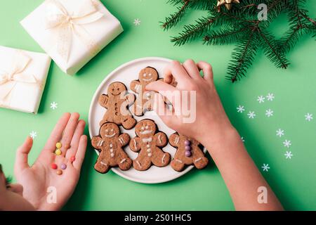 Blick von oben auf die Hände, die Lebkuchenkekse mit festlicher Dekoration, gewickelten Geschenken und Tannenzweigen auf grünem Hintergrund dekorieren. Stockfoto