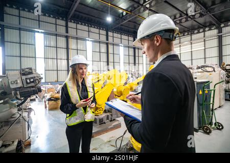 Zwei Fachleute mit Schutzhelm nehmen an einer Diskussion in einer Produktionsstätte Teil. Einer hält eine Zwischenablage, während der andere Beobachtungen feststellt, Stockfoto
