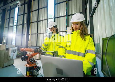 Zwei Arbeiter in hellen Sicherheitswesten und Helmen interagieren in einer industriellen Umgebung mit einem Laptop. Ein Roboterarm ist sichtbar und hebt das moderne Au hervor Stockfoto