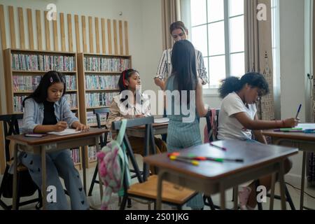 Kinder nehmen an einem Kunstunterricht Teil, wobei sie mit farbigen Bleistiften und Papier arbeiten, während ein Lehrer sie in einem hellen und organisierten Klassenzimmer mit bo führt Stockfoto