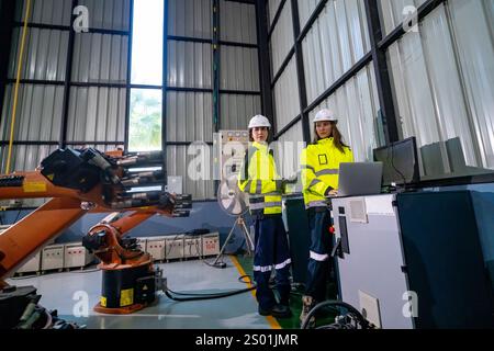 Zwei Ingenieure in Sicherheitsausrüstung stehen neben einem Roboterarm in einem hellen Industriebereich. Sie überwachen den Betrieb und besprechen automatisierte Prozesse Stockfoto