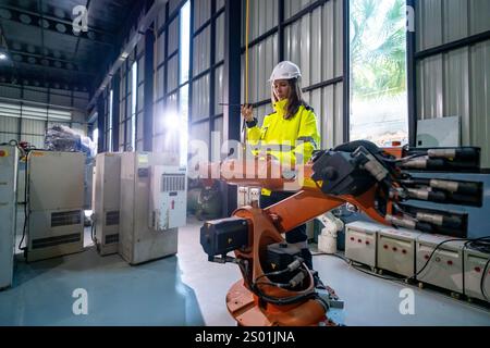 Eine Ingenieurin in Sicherheitsausrüstung inspiziert einen Roboterarm in einer gut beleuchteten Industrieanlage. Natürliches Licht filtert durch die Fenster und beleuchtet die Sonne Stockfoto