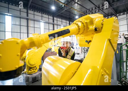 Ein Ingenieur mit Schutzhelm verwendet ein Funkgerät, um zu kommunizieren, während er einen gelben Roboterarm bei der Arbeit in einer High-Tech-Fertigungsanlage beobachtet und Auto präsentiert Stockfoto