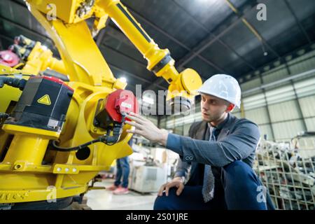 Ein Arbeiter mit Schutzhelm untersucht einen gelben Roboterarm genau. Die Umgebung ist eine geräumige Produktionsstätte mit verschiedenen Maschinen im Hintergrund Stockfoto