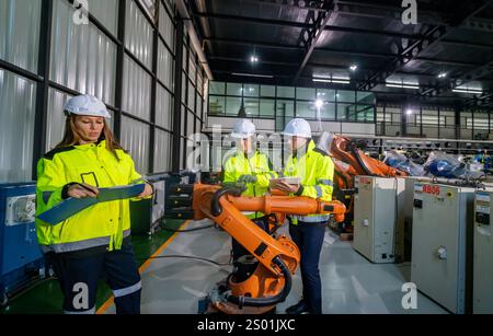 Drei Ingenieure besprechen einen Roboterarm, während sie Daten auf Tablets überprüfen. Sie sind in Sicherheitsausrüstung gekleidet und arbeiten in einer geräumigen, gut beleuchteten Industrie Stockfoto