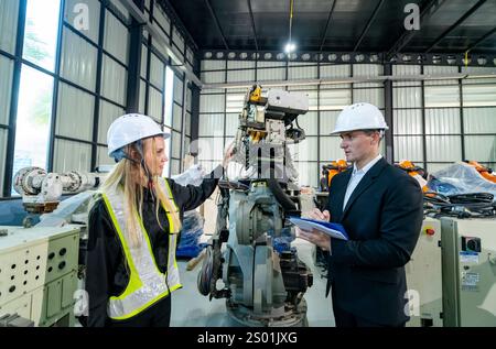 In einer modernen Industrieanlage erklärt ein Techniker in Sicherheitsausrüstung einem Ingenieur die Funktionsweise eines Roboterarms. Beide Individuen konzentrieren sich auf mA Stockfoto
