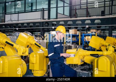 Ein Techniker mit gelbem Schutzhelm sitzt zwischen gelben Roboterarmen und arbeitet in einer modernen Industrieanlage mit Computern. Die helle Arbeit Stockfoto