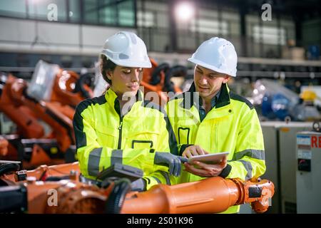 Zwei Ingenieure, die Schutzhelme und helle Jacken tragen, diskutieren über ein Robotikprojekt in einer großen Industrieanlage. Stockfoto