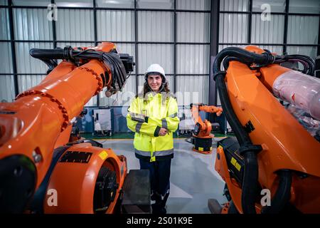 Ein Profi in einer Sicherheitsjacke und einem Helm posiert stolz zwischen zwei industriellen Roboterarmen in einem technologiegesteuerten Arbeitsbereich. Die helle Umgebung Stockfoto