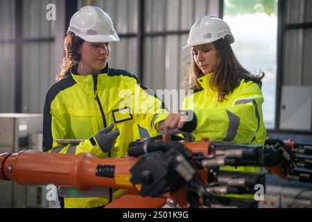 In Sicherheitsausrüstung gekleidete Ingenieure arbeiten gemeinsam an einem Roboterarm in einem Fertigungswerk. Sie sind fokussiert, besprechen Details und stellen sicher, dass sie professionell sind Stockfoto