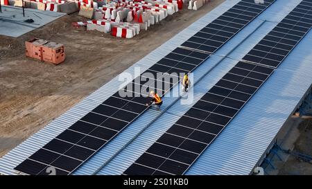 Zwei Arbeiter werden beobachtet, wie sie Solarpaneele auf einem Welldach sorgfältig installieren. Der Standort wird derzeit gebaut, wobei die Materialien in der verstreut sind Stockfoto