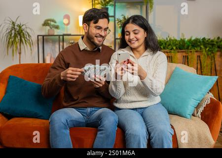 Glückliches indisches Paar, das Geld hält, Bargeld und Smartphone plant, zusammen über Investitionen zu diskutieren, sitzend auf Sofa im Wohnzimmer zu Hause. Erfolgreiche, vielfältige Familie auf Couch in der Wohnung. Stockfoto
