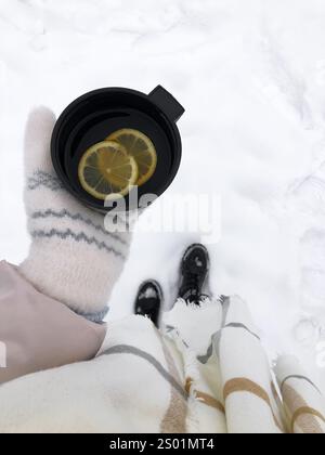 Eine gemütliche Winterszene mit Handschuhen, die eine schwarze Tasse warmen Tee halten, mit zwei hellen Zitronenscheiben auf der Oberfläche. Die schneebedeckte gr Stockfoto