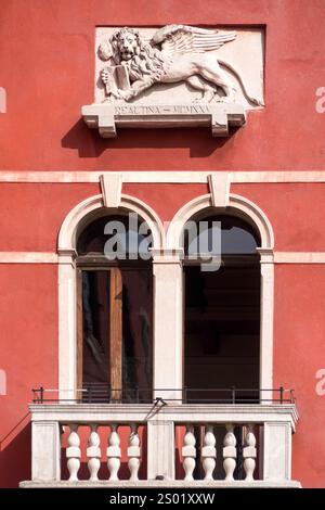 VENEDIG, ITALIEN - 06. MAI 2015: Tafel mit geflügeltem Löwen auf einer roten Gipsmauer über Fenster und Balkon Stockfoto