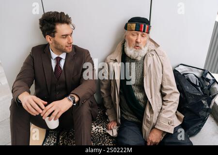 Netter junger Geschäftsmann im Anzug, der der Geschichte eines älteren Obdachlosen lauscht, der auf dem Bürgersteig sitzt und Mitgefühl und Empathie für weniger glückliche Menschen zeigt. Stockfoto
