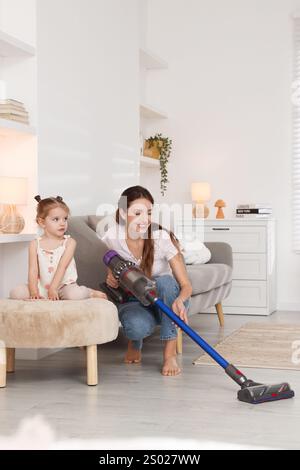 Die lächelnde junge Frau und ihre Tochter putzen zu Hause den Boden mit schnurlosem Staubsauger Stockfoto