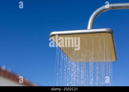 Quadratischer Duschkopf im Freien mit fließendem Wasser, erfasst vor einem klaren blauen Himmel und Teil einer Dachkonstruktion. Stockfoto