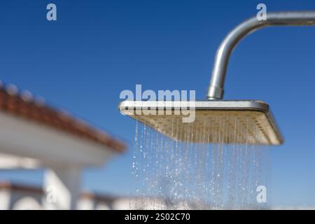 Nahaufnahme einer quadratischen Außendusche mit Wassertropfen, die vor einem klaren blauen Himmel und einer verschwommenen Dachterrasse liegen. Stockfoto