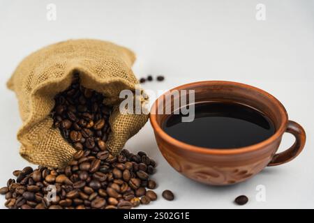 Kaffeebohnen, die aus einem Sacksack verschüttet werden, neben einer Tontasse gefüllt mit schwarzem Kaffee auf weißem Hintergrund, rustikaler und natürlicher Stil. Stockfoto