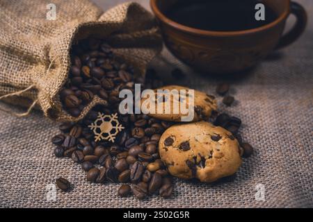 Rustikales Stillleben mit einem Sack, der Kaffeebohnen, Schokoladenchips-Cookies und einer Keramikschale Kaffee auf einem Sackleinen-Hintergrund verschüttet. Stockfoto