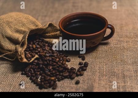 Eine braune Keramikschale gefüllt mit schwarzem Kaffee neben einem Sack, der geröstete Kaffeebohnen auf eine strukturierte Sackfläche verschüttet. Stockfoto