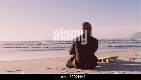 Afroamerikaner sitzt ruhig am Strand, mit Kopierraum. Er trägt einen Neoprenanzug und schlägt eine Pause in seiner Surfsession vor, um nachzudenken. Stockfoto