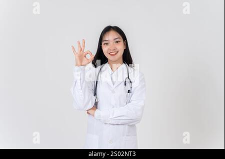 Eine positive, lächelnde asiatische Ärztin in einer weißen Uniform, die das OK-Handzeichen vor der Kamera zeigt, steht vor einem weißen Studiohintergrund. p Stockfoto