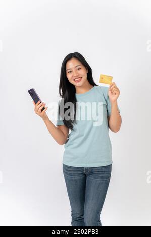 Eine attraktive, lächelnde asiatische Frau in einem blauen T-Shirt, die vor weißem Studiohintergrund steht und ein Smartphone und eine Kreditkarte hält. Mobile Bank Stockfoto