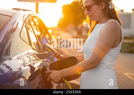 Eine Frau in einem weißen Kleid benutzt ein Handy, während sie ihr Elektroauto während Sonnenuntergang auflädt Stockfoto