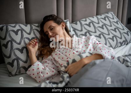 Eine Frau im Schlafanzug mit Herzmuster schläft ruhig im Bett, eingebettet zwischen Kissen und Decken. Eine ruhige Morgenszene. Stockfoto
