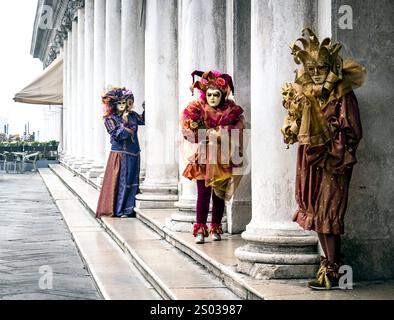 Drei Personen in aufwendigen Kostümen und Masken stehen neben klassischen Säulen. Die Umgebung scheint ein historischer oder kultureller Ort zu sein Stockfoto