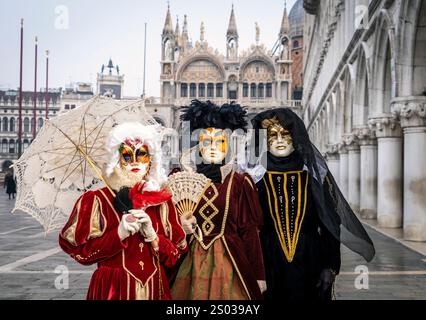 Drei Personen in aufwendigen Kostümen und Masken stehen auf einem historischen Platz mit einem Ventilator und einem Regenschirm. Die Kulisse bietet eine kunstvolle Architektur Stockfoto
