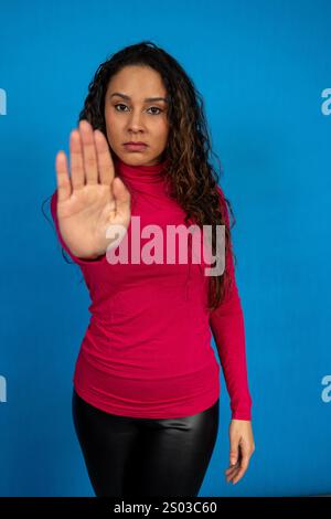 Studio-Porträt einer ernsten jungen Frau, die mit ihrer Hand ein Stoppschild macht, isoliert auf blauem Hintergrund Stockfoto