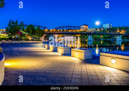 Ein Stadtpark mit einer Brücke und einem Fluss. Die Brücke ist nachts beleuchtet. Der Mond ist am Himmel sichtbar Stockfoto
