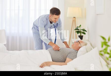 Älterer alter Mann, der zu Hause im Bett liegt, junger Arzt, männliche Krankenschwester, die sich um den alten Patienten kümmert Stockfoto
